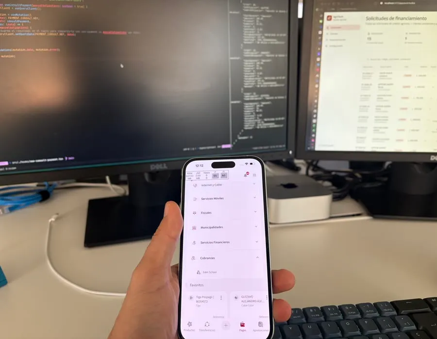 Mano sosteniendo un teléfono con una app de pagos hecha en React Native, y el código detrás en los monitores